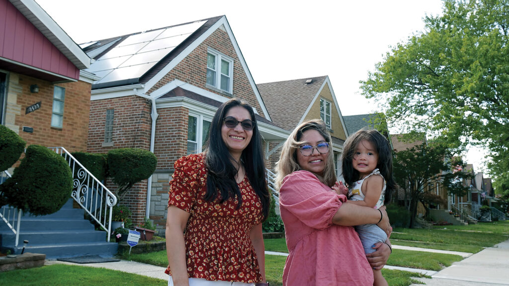 Hispanic family pose in front of a brick-house with solar panels: a woman with dark glasses and long hair, a second woman with windswept gray hair holding a young child with a full head of long brown hair.